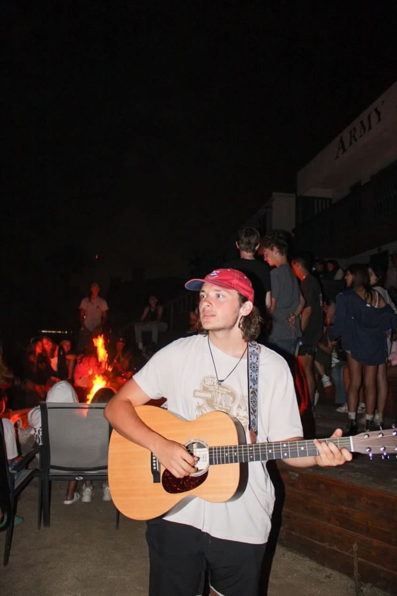 Colby playing acoustic guitar beside a bonfire at night, a crowd gathered behind him